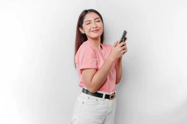 幸せなアジアの女性の肖像画は ピンクのTシャツを着て 白い背景によって隔離された彼女の携帯電話を保持している — ストック写真