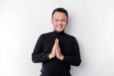 Smiling young Asian man wearing black shirt, gesturing traditional greeting isolated over white background