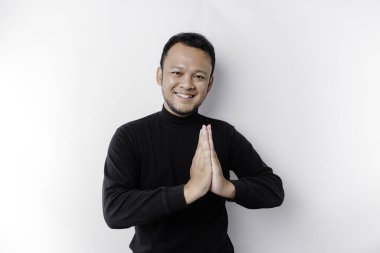 Smiling young Asian man wearing black shirt, gesturing traditional greeting isolated over white background