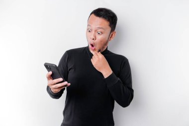 Şok olmuş, siyah tişörtlü, ağzı açık, beyaz arka planla izole edilmiş, telefonunu tutan Asyalı bir adam.