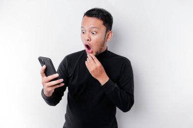 Şok olmuş, siyah tişörtlü, ağzı açık, beyaz arka planla izole edilmiş, telefonunu tutan Asyalı bir adam.