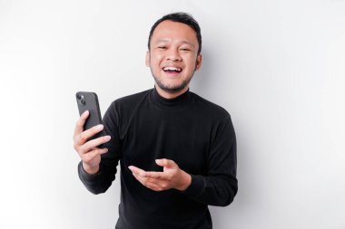 Siyah tişörtlü heyecanlı Asyalı adam telefonunu tutarken gülümsüyor, beyaz arka planla izole edilmiş.