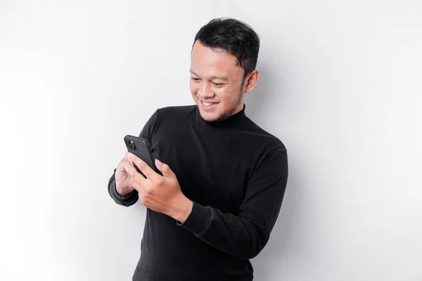 stock image Excited Asian man wearing black shirt smiling while holding his phone, isolated by white background