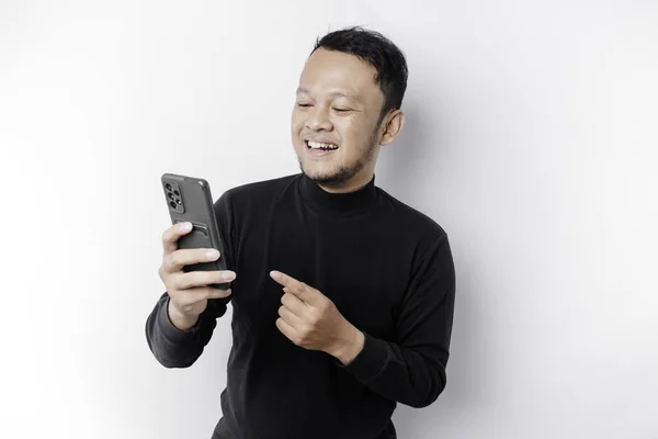 stock image Excited Asian man wearing black shirt smiling while holding his phone, isolated by white background