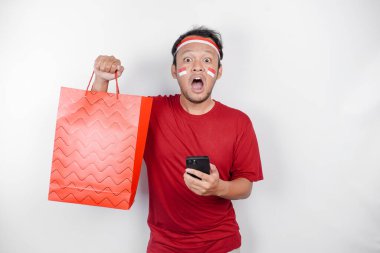Çekici bir Asyalı elinde online alışveriş çantası ve beyaz arka planda izole edilmiş akıllı telefonu ile heyecanla bekliyor. Endonezya 'nın Bağımsızlık Günü konsepti