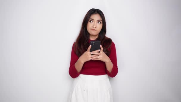 思慮深い若いアジアの女性が赤いTシャツを着て 彼女の携帯電話をホワイトバックグラウンドで孤立したアイデアに触発された見た目を保持しています — ストック動画