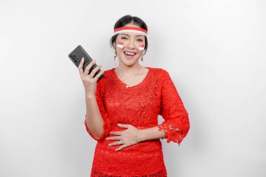 Kırmızı kebaya ve saç bandı takmış düşünceli bir Asyalı kadın, telefonu ve karnını tutuyor, ne yiyeceğini ya da ne sipariş edeceğini düşünüyor, beyaz arka planla izole edilmiş. Endonezya 'nın bağımsızlık günü