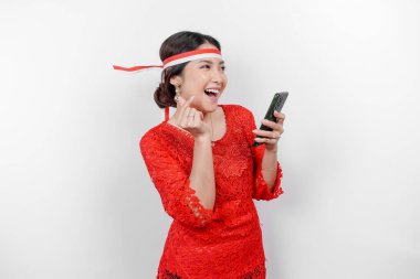 Kırmızı kebaya ve saç bandı takan mutlu bir Asyalı kadın, telefonunu tutuyor ve beyaz arka planla izole edilmiş, şefkatli duygularını gösteren parmaklarıyla işaret ediyor. Endonezya 'nın bağımsızlık günü