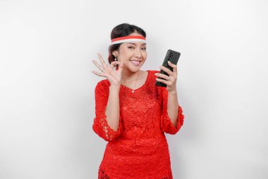 Kırmızı kebaya ve saç bandı takan neşeli bir Asyalı kadın, beyaz arka planla izole edilmiş parmaklarıyla işaret ederken telefonunu tutuyor. Endonezya 'nın bağımsızlık günü konsepti