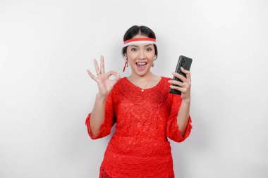 Kırmızı kebaya ve saç bandı takan neşeli bir Asyalı kadın, beyaz arka planla izole edilmiş parmaklarıyla işaret ederken telefonunu tutuyor. Endonezya 'nın bağımsızlık günü konsepti