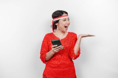 Kırmızı kebaya ve saç bandı takan mutlu bir Asyalı kadın, telefonunu tutuyor ve beyaz arka planla izole edilmiş fotokopi alanını ona doğrultuyor. Endonezya 'nın bağımsızlık günü