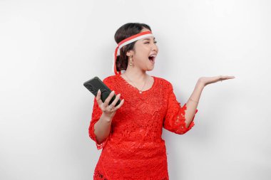 Kırmızı kebaya ve saç bandı takan mutlu bir Asyalı kadın, telefonunu tutuyor ve beyaz arka planla izole edilmiş fotokopi alanını ona doğrultuyor. Endonezya 'nın bağımsızlık günü
