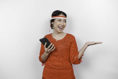 Kırmızı kebaya ve saç bandı takan mutlu bir Asyalı kadın, telefonunu tutuyor ve beyaz arka planla izole edilmiş fotokopi alanını ona doğrultuyor. Endonezya 'nın bağımsızlık günü