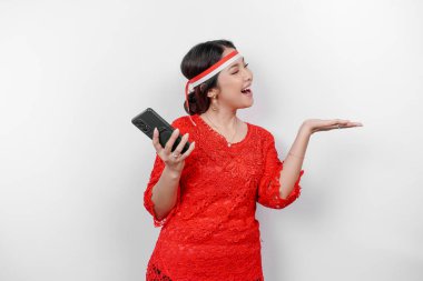 Kırmızı kebaya ve saç bandı takan mutlu bir Asyalı kadın, telefonunu tutuyor ve beyaz arka planla izole edilmiş fotokopi alanını ona doğrultuyor. Endonezya 'nın bağımsızlık günü