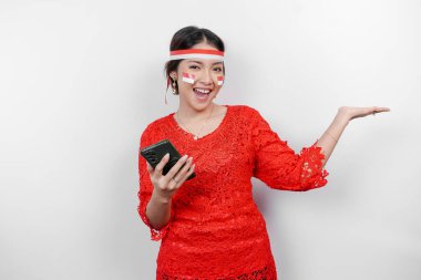 Kırmızı kebaya ve saç bandı takan mutlu bir Asyalı kadın, telefonunu tutuyor ve beyaz arka planla izole edilmiş fotokopi alanını ona doğrultuyor. Endonezya 'nın bağımsızlık günü