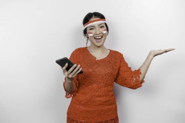 Kırmızı kebaya ve saç bandı takan mutlu bir Asyalı kadın, telefonunu tutuyor ve beyaz arka planla izole edilmiş fotokopi alanını ona doğrultuyor. Endonezya 'nın bağımsızlık günü