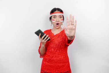 Kırmızı kebaya takan çekici Asyalı kadın ve el hareketleriyle saç bandı. Beyaz arka planla izole edilmiş telefonunu tutarken reddedilme ya da yasaklama pozu veriyor.
