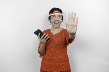 Kırmızı kebaya takan çekici Asyalı kadın ve el hareketleriyle saç bandı. Beyaz arka planla izole edilmiş telefonunu tutarken reddedilme ya da yasaklama pozu veriyor.