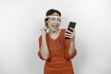 Telefonunu tutarken ve kırmızı kebaya takarken mutlu ve başarılı bir ifadeye sahip genç bir Asyalı kadın, beyaz arka planla izole edilmiş bayrak bandı. Endonezya 'nın bağımsızlık günü konsepti.