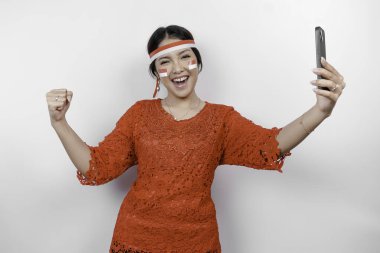 Telefonunu tutarken ve kırmızı kebaya takarken mutlu ve başarılı bir ifadeye sahip genç bir Asyalı kadın, beyaz arka planla izole edilmiş bayrak bandı. Endonezya 'nın bağımsızlık günü konsepti.