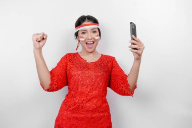 Telefonunu tutarken ve kırmızı kebaya takarken mutlu ve başarılı bir ifadeye sahip genç bir Asyalı kadın, beyaz arka planla izole edilmiş bayrak bandı. Endonezya 'nın bağımsızlık günü konsepti.