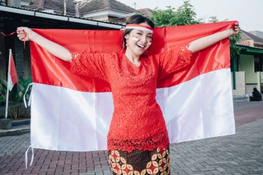 Endonezya Bağımsızlık Günü 'nü kutlamak için elinde Endonezya bayrağı tutan kırmızı kebaya giymiş mutlu Endonezyalı kadın. Açık hava fotoşopu kavramı