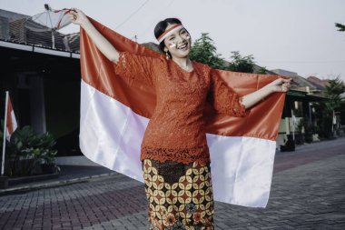 Endonezya Bağımsızlık Günü 'nü kutlamak için elinde Endonezya bayrağı tutan kırmızı kebaya giymiş mutlu Endonezyalı kadın. Açık hava fotoşopu kavramı