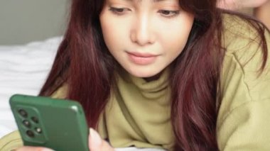 Genç bir Asyalı kadın kendini ciddi hissediyor ve yatakta yatarken akıllı telefonunu elinde tutmaya odaklanmış durumda..