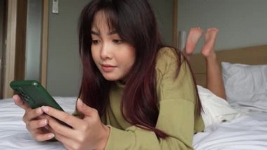 Genç bir Asyalı kadın kendini ciddi hissediyor ve yatakta yatarken akıllı telefonunu elinde tutmaya odaklanmış durumda..