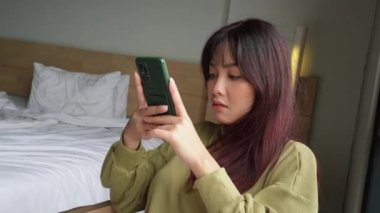 Genç bir Asyalı kadın kendini ciddi hissediyor ve yatak odasında otururken akıllı telefonunu elinde tutmaya odaklanıyor..