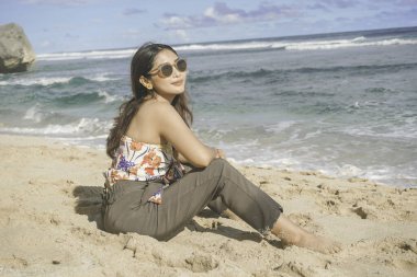 Genç Asyalı kadın kumsalda oturuyor. Portre seksi Asyalı bayan yazın tropikal doğayla seyahat ediyor ve dinleniyor..