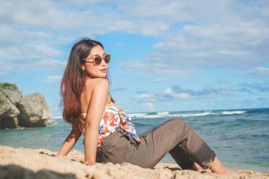 Genç Asyalı kadın kumsalda oturuyor. Portre seksi Asyalı bayan yazın tropikal doğayla seyahat ediyor ve dinleniyor..