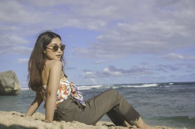 Genç Asyalı kadın kumsalda oturuyor. Portre seksi Asyalı bayan yazın tropikal doğayla seyahat ediyor ve dinleniyor..