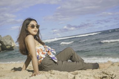 Genç Asyalı kadın kumsalda oturuyor. Portre seksi Asyalı bayan yazın tropikal doğayla seyahat ediyor ve dinleniyor..