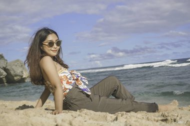 Genç Asyalı kadın kumsalda oturuyor. Portre seksi Asyalı bayan yazın tropikal doğayla seyahat ediyor ve dinleniyor..