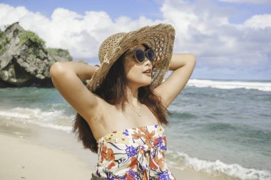 Plaj şapkası takan mutlu bir Asyalı kadın elleri açık kameraya poz veriyor. Gunungkidul, Endonezya sahilinde özgürlüğünü ve tasasız tatilini ifade ediyor.