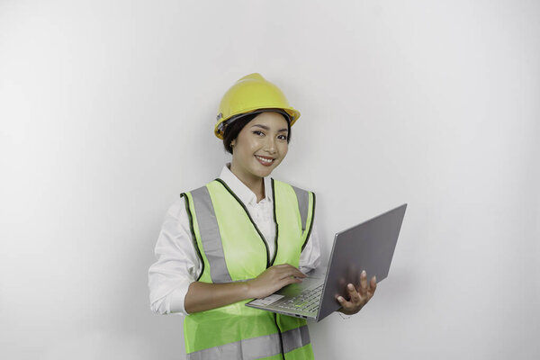 A smiling Asian woman labor wearing safety helmet and vest while holding her laptop, isolated by white background. Labor's day concept.