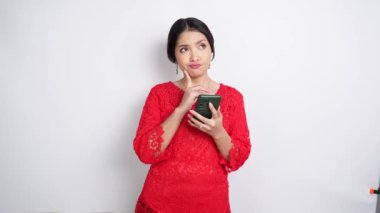 Saç bandı ve kırmızı kebaya takan zeki genç bir kadın beyaz arka planla izole edilmiş telefonunu tutarken fotokopi uzayı ile ilgili bir fikre bakıyor. Endonezya 'nın bağımsızlık günü.