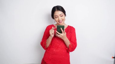 Kırmızı kebaya ve saç bandı takmış gülümseyen Asyalı bir kadın ve beyaz arka planla izole edilmiş telefonunu tutuyor. Endonezya 'nın bağımsızlık günü konsepti