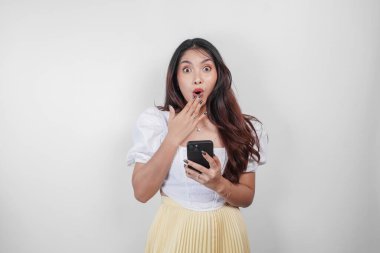 Beyaz arka planla izole edilmiş akıllı telefonunu tutarken ağzı açık, şok olmuş Asyalı bir kadın..