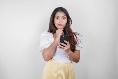 Düşünceli bir Asyalı kadın, beyaz arka planla izole edilmiş düşüncelerini hayal ederken akıllı telefonunu tutuyor..