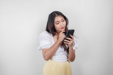 Düşünceli bir Asyalı kadın, beyaz arka planla izole edilmiş düşüncelerini hayal ederken akıllı telefonunu tutuyor..