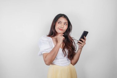 Düşünceli bir Asyalı kadın, beyaz arka planla izole edilmiş düşüncelerini hayal ederken akıllı telefonunu tutuyor..