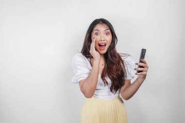 Genç ve güzel bir Asyalı kadın, beyaz arka planla izole edilmiş telefonunu tutarken eli ağzında bağırıyor ve çığlık atıyor..