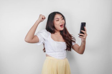 Genç ve güzel bir Asyalı kadın beyaz arka planla izole edilmiş telefonunu tutarken tezahürat yapıyor..