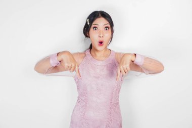 Shocked Asian woman dressed in pink, pointing at the copy space below her, isolated by white background