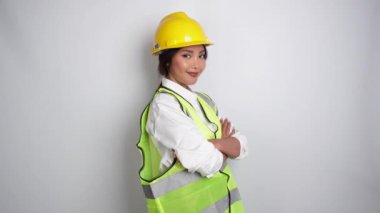 Gülümseyen Asyalı kadın işçi sanayi fabrikasında, kolları bağlı poz veriyor, sarı kasklı, yeşil yelekli ve üniformalı, izole beyaz arka planda..