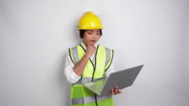 Dizüstü bilgisayarını ve elini beyaz arka planda tutarken çelik yelek ve miğfer giyen düşünceli genç bir işçi. İşçi Bayramı konsepti.