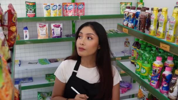 一位亚洲女人拿着平板电脑 检查商店的存货 一位出纳员穿着黑色围裙走在杂货店或便利店的过道上 — 图库视频影像