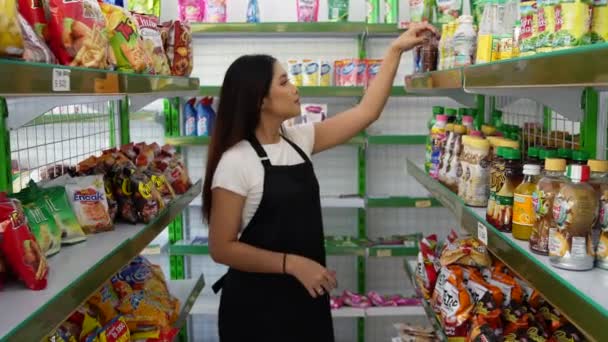 一位亚洲妇女正在检查和整理商店的存货 一位出纳员穿着黑色围裙走在杂货店或便利店的过道上 — 图库视频影像